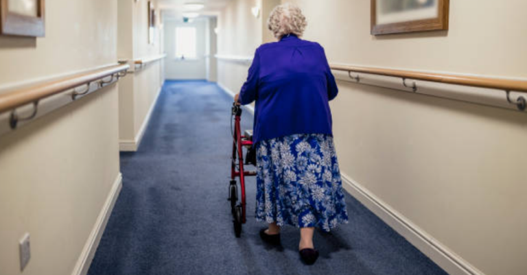 An elderly woman using a walker to navigate down a well-lit corridor, appearing safe and independent.