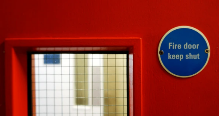 Image of a closed red fire door equipped with safety signage and a push bar, demonstrating compliance with fire safety regulations in a nursing home setting.