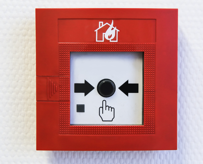 A red manual call point (MCP) for a fire alarm system, featuring a central black button with arrows pointing towards it and an icon of a hand pressing the button, with a fire symbol above.