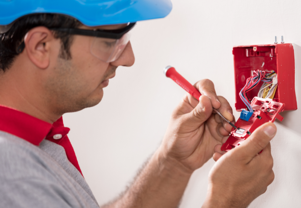 Technician working on a fire alarm system, ensuring compliance with fire safety regulations as part of the Nursing Home Resident Safety Improvement Scheme in Ireland.