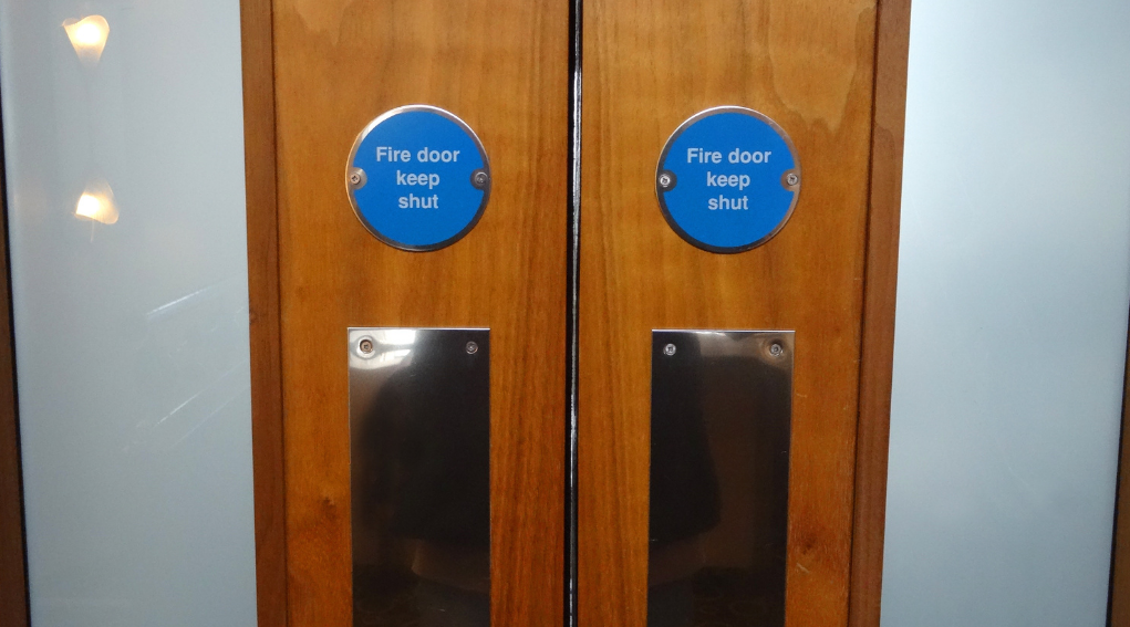 Two adjacent wooden fire doors with circular blue “Fire door keep shut” signs and metal push plates, illustrating typical safety signage and hardware on fire-rated doors.